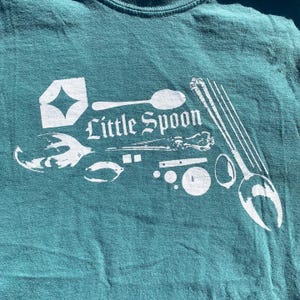 May include: A teal t-shirt featuring a white graphic print of various kitchen utensils, including a spoon, a fork, and a whisk, alongside the text "Little Spoon" in a stylised font. The design also includes small geometric shapes and circles.