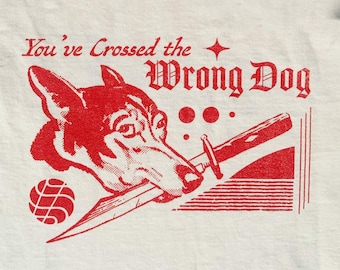 May include: An off-white t-shirt featuring a red graphic print of a dog holding a dagger in its mouth. The text reads, "You've Crossed the Wrong Dog." The design includes a small four-pointed star, three circular dots, and a circular geometric emblem.