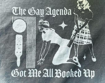 Peut inclure: Un t-shirt gris foncé avec une impression graphique blanche. Le texte indique The Gay Agenda en haut et Got Me All Booked Up en bas. L'illustration représente une personne à genoux, une personne debout, une forme circulaire et une raquette.