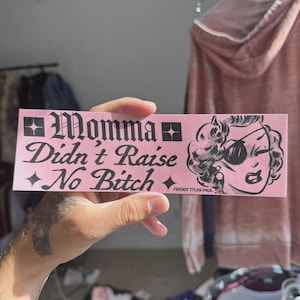 May include: A pink sticker with the text "Momma Didn't Raise No Bitch" in a gothic font. The sticker also features a cartoon illustration of a woman wearing sunglasses. The artist's name, "FREDDY TYLER PAUL," is printed at the bottom.