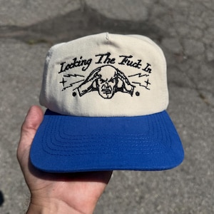 May include: A cream-colored baseball cap with a blue brim. The front panel features black embroidered text that reads "Locking The Fuck In" above a graphic illustration of a person holding their head with lightning bolts on either side.