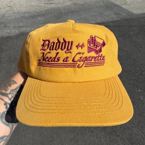 Pode incluir: Um boné de beisebol amarelo-mostarda com bordado roxo escuro na parte frontal. O texto "Daddy Needs a Cigarette" está escrito numa fonte estilizada, acompanhado por um pequeno gráfico bordado de um maço de cigarros e duas estrelas.