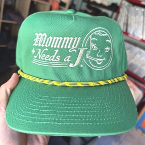 May include: Green trucker hat with the embroidered text "Mommy Needs a J." in white script. The hat features a yellow and black rope detail and a cartoon face graphic. A classic, casual accessory.