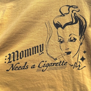 May include: Mustard yellow t-shirt featuring the text "Mommy Needs a Cigarette" and a black illustration of a woman smoking. The woman has dark hair and an earring. Two black diamond shapes are to the right of the woman's head.