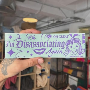 Puede incluir: Una pegatina rectangular con fondo verde claro y texto e ilustraciones moradas. El texto dice "I'm Dissociating Again" en una fuente gótica, con las palabras "OH GREAT" encima. La pegatina también presenta ilustraciones de ojos y boca, y un dibujo de una niña.