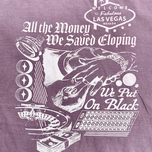 May include: A purple t-shirt with a white graphic design. The graphic features a pair of hands holding a ring, a roulette wheel, and the text "All the Money We Saved Eloping We Put On Black". The graphic also includes a sign that says "Welcome to Fabulous Las Vegas, Nevada".