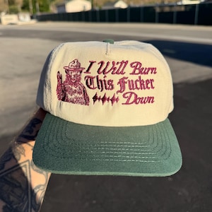 May include: A beige baseball cap with a dark green brim. The front features an embroidered graphic of a bear wearing a ranger hat, accompanied by the maroon text "I Will Burn This Fucker Down" with decorative diamond-shaped accents between the words.