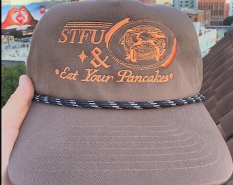 May include: A brown baseball cap with the embroidered text "STFU & Eat Your Pancakes" in orange. The cap features a circular design with a pancake illustration and a black and white rope detail.