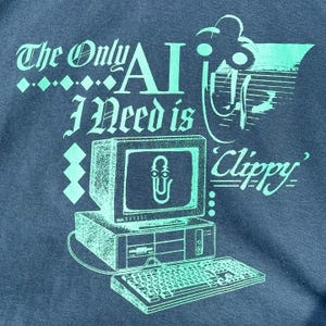 Op de afbeelding: Een blauw t-shirt met een afbeelding van een vintage computer met de tekst "The Only AI I Need is Clippy".