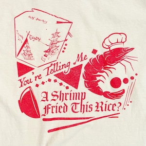 May include: A cream-coloured t-shirt featuring a red graphic print of a takeaway box, a shrimp wearing a chef hat, and a bowl. The text reads: You're Telling Me A Shrimp Fried This Rice? The design includes geometric shapes and lines.