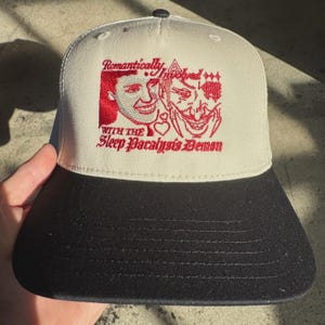 Könnte beinhalten: Beige und schwarze Baseballkappe mit dem roten Text "Romantically Involved With The Sleep Paralysis Demon". Die Kappe hat ein gesticktes Design von zwei Gesichtern und anderen Symbolen.