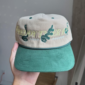 Pode incluir: Boné de beisebol bege e verde com o texto "FREDDY TIGER IC" bordado em verde claro. O boné tem uma aba verde e um detalhe de corda verde. Duas cobras verdes bordadas estão no boné.