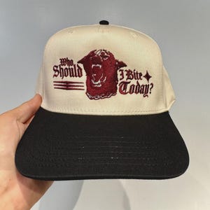 Puede incluir: Gorra de béisbol beige y negra con el texto "Who Should I Bite Today?" bordado en borgoña. La gorra presenta un gráfico de animal gruñendo y una visera plana. La gorra tiene un botón negro en la parte superior.