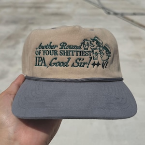 May include: Tan and gray baseball cap with the embroidered text "Another Round of Your Shittiest IPA, Good Sir!" and a graphic of two men drinking beer. The cap has a gray brim and a rope detail.