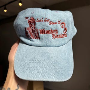 Può includere: Un cappello da baseball in denim blu chiaro con testo ricamato in rosso che recita "OK Let's Get Down To Monkey Business". Il ricamo include anche un'immagine di un uomo in costume.
