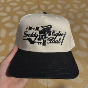 May include: A white and black baseball cap with the text "Freddy Eyler Paul" and a panther graphic. The cap has a flat brim and a structured crown. The design is in black.