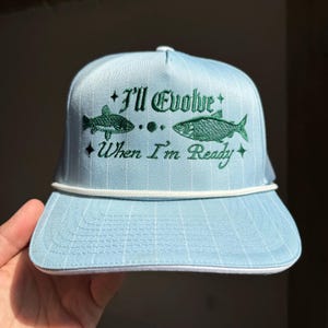 May include: A light blue baseball cap with a white brim and a green embroidered design of two fish and the text "I'll Evolve When I'm Ready".