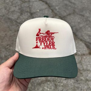 Może przedstawiać: Biało-zielona czapka z daszkiem z haftowanym na czerwono logo z napisem "Freddy Tyler Paul" i grafiką kobiety trzymającej karabin.