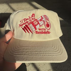 May include: A beige baseball cap with a red embroidered design. The design includes the text "I Don't Negotiate With Bombshells" and an image of a woman, a bomb, and a clock. The cap has a curved brim and a structured crown.