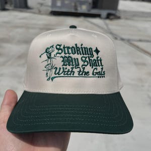 Puede incluir: Gorra de béisbol beige y verde con el texto "Stroking My Shaft With the Gals" bordado en verde. La gorra tiene una visera verde y un diseño temático de golf.