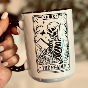 Puede incluir: Taza de cerámica blanca con asa y borde negros. La taza presenta una ilustración en blanco y negro de un esqueleto leyendo un libro, con el texto "THE READER" debajo. La taza forma parte de la colección Graveyard & Co.