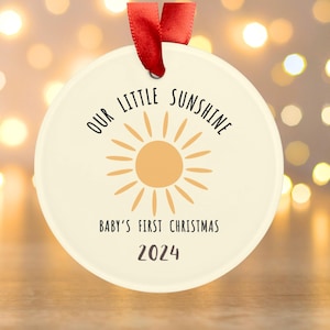 May include: A white round ornament with a yellow sun and the text "Our Little Sunshine Baby's First Christmas 2024".