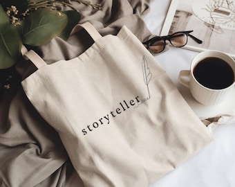Puede incluir: Una bolsa de tela beige con la palabra storyteller impresa en letras minúsculas negras, junto a un dibujo lineal negro de una pluma de escribir que se extiende desde la letra r. La bolsa cuenta con dos asas de tela a juego.