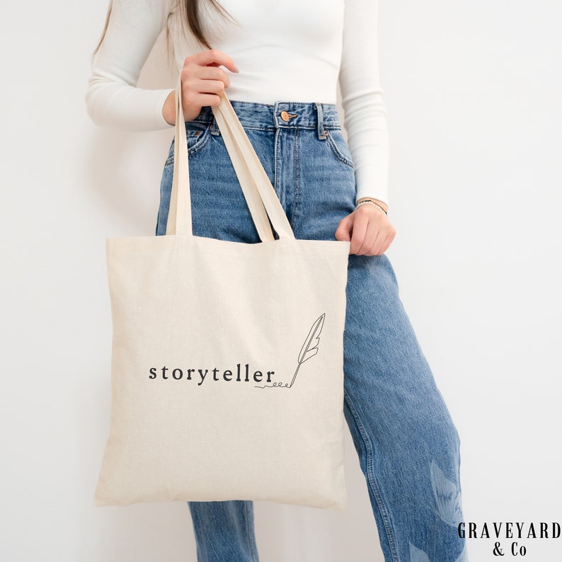 May include: A white canvas tote bag with black text that reads "storyteller" and a simple line drawing of a quill pen.