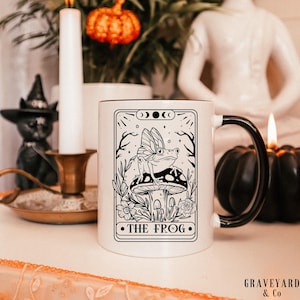 May include: A white ceramic mug with a black handle featuring a black line art illustration of a frog with wings perched on a mushroom. The design is framed like a tarot card with the text THE FROG at the bottom, surrounded by floral motifs.