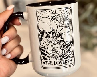 May include: A white ceramic mug with a black interior and handle, featuring a black line art illustration of two skulls facing each other, surrounded by roses and stars, with the Roman numeral VI and the text THE LOVERS printed below.