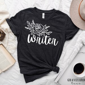 May include: A dark gray t-shirt with a white floral design and the word "writer" in white cursive lettering.