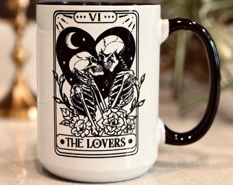 May include: A white ceramic mug with a black handle and rim, featuring a black graphic print of two skeletons embracing inside a heart shape. The design resembles a tarot card labeled VI and THE LOVERS, surrounded by floral accents.