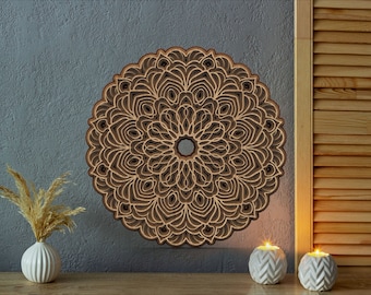 Könnte beinhalten: Ein mehrschichtiges Wandkunstwerk mit Mandala-Motiv aus Holz mit einem detaillierten, symmetrischen Design. Das Kunstwerk ist in Braun- und Beigetönen gehalten und an einer grau strukturierten Wand befestigt. Dekorative Kerzen und eine Vase mit getrockneten Pflanzen sind im Vordergrund.