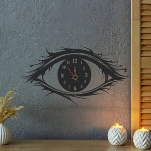 May include: A black wall clock in the shape of an eye, with a clock face at the center. The clock has black numbers and hands, with a red second hand. The clock is mounted on a gray textured wall. Decorative candles and a vase are in the foreground.