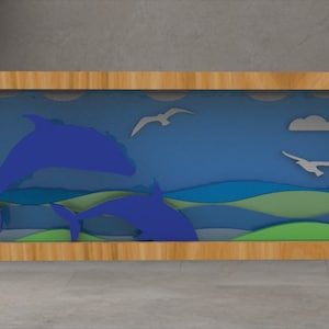 May include: A framed artwork featuring a layered ocean scene with blue dolphins, waves, clouds, and seagulls. The artwork is set against a blue sky and green and blue waves, framed in a light wood.