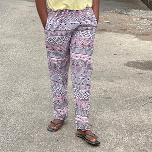 May include: A woman wearing a yellow halter top and patterned pants stands on a paved road. The pants feature a geometric design in black, white, and red. She is smiling, with her hands in her pockets, and wearing sandals.