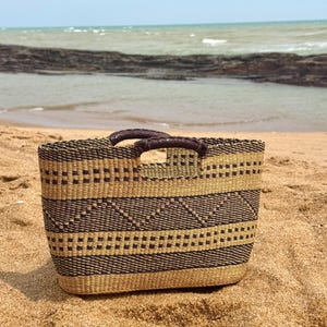 Könnte beinhalten: Ein rechteckiger Webkorb mit braunen und beigefarbenen geometrischen Mustern und zwei braunen Ledergriffen. Der Korb steht an einem Sandstrand mit dem Meer im Hintergrund.
