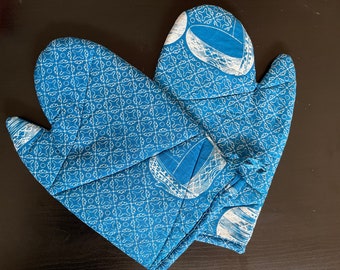 May include: A pair of blue oven mitts with a white pattern. The mitts feature a quilted design with a repeating geometric and circular motif. The design includes a white illustration of a bowl. The oven mitts are designed for kitchen use.