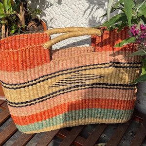 May include: A rectangular woven basket bag featuring horizontal stripes in shades of orange, yellow, light green, and black. The bag has two integrated woven handles at the top and a structured, open-top design with a textured, fibrous surface.