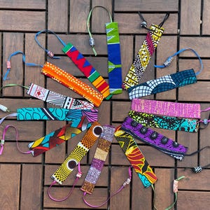 May include: A collection of colorful fabric bookmarks with patterned designs, arranged in a circular pattern. Each bookmark has a ribbon attached with beads. The bookmarks feature various colors and patterns, including geometric and floral motifs.