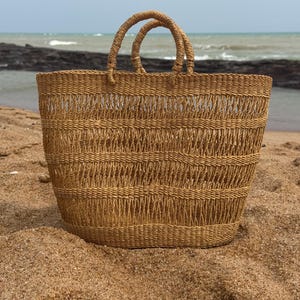 May include: A tan woven straw tote bag featuring an open-weave design with horizontal bands of tighter stitching and two rounded top handles. The bag has a wide, rectangular base and a structured body, showcasing a natural light brown colour.