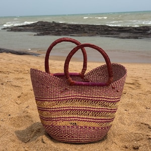 Könnte beinhalten: Eine geflochtene Korbtasche mit einem Streifenmuster in Dunkelburgund und Metallic-Gold. Die Tasche hat einen abgerundeten Boden und zwei stabile, passende burgunderrote kreisförmige Griffe am oberen Rand, die ein strukturiertes Design bilden.