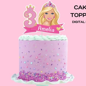 Op de afbeelding: Roze en gouden taarttopper met een Barbiepop met een kroon en het nummer 3. De topper bevat ook een banner met de naam Amelia.