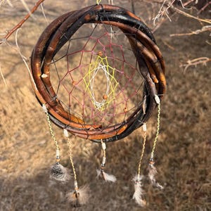 May include: A handmade dreamcatcher with a circular frame woven from brown branches. The web inside is a mix of red, yellow, and white threads, with small beads. Feathers and beads hang from the bottom, suspended by gold-coloured chains.