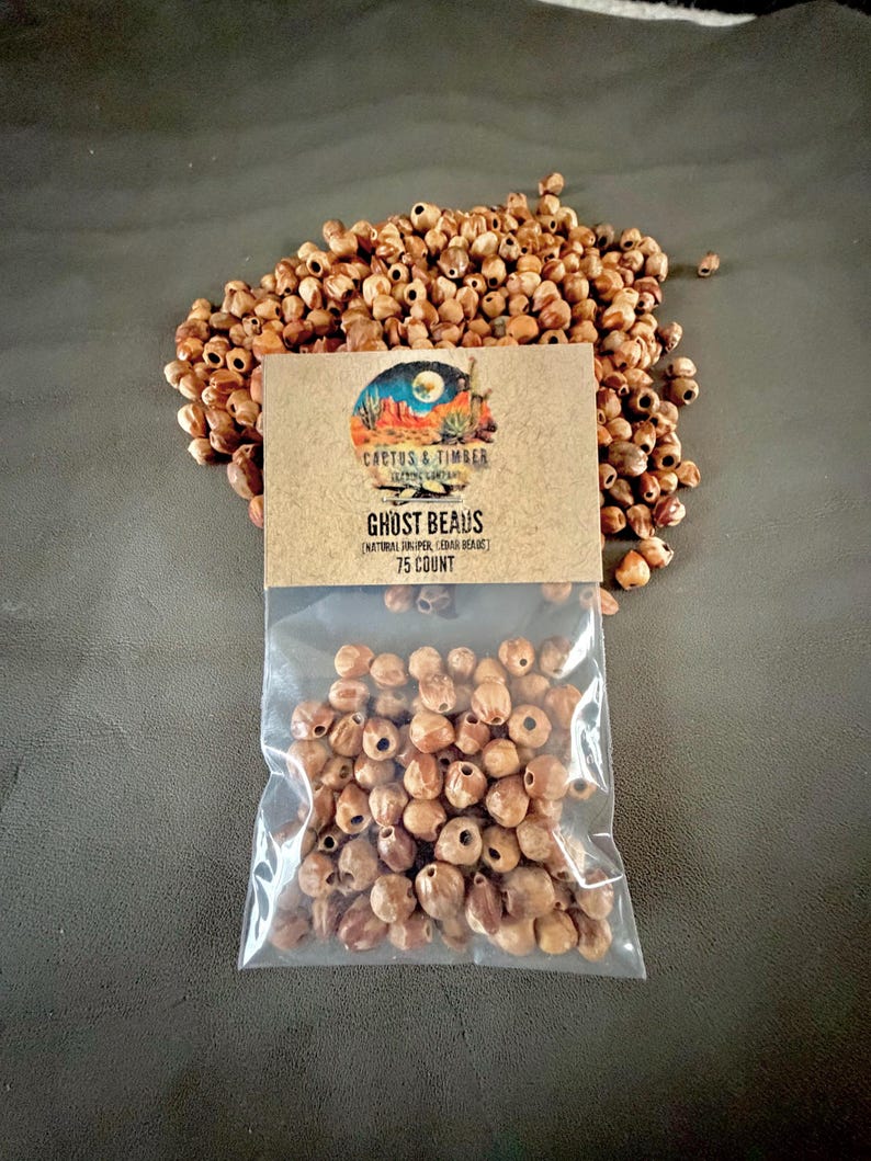 May include: A clear plastic bag containing 75 natural juniper cedar beads, with a tan paper label featuring the text Cactus & Timber, Ghost Beads, and Natural Juniper Cedar Beads, placed in front of a loose pile of identical brown wooden beads.