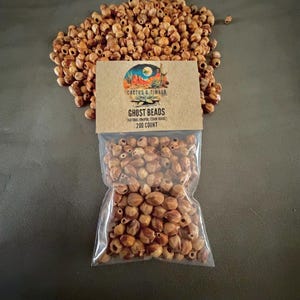 May include: A collection of small, light brown, natural juniper cedar beads, with a portion loose and a portion contained in a clear plastic bag. The bag features a tan paper header with the text Cactus & Timber, Ghost Beads, and 200 count.