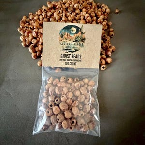 May include: A pile of small, light brown, round natural juniper cedar beads with a hole in the center. A clear plastic bag contains a portion of the beads, topped with a tan paper label reading Cactus & Timber Trading Company Ghost Beads.