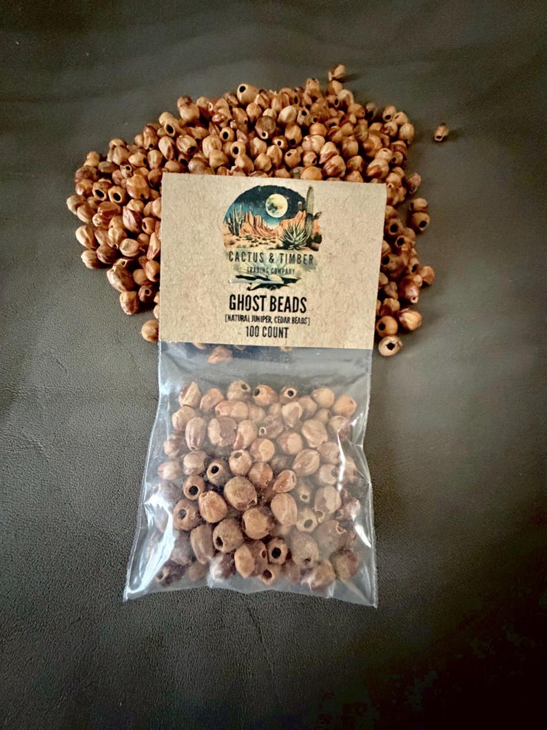 May include: A pile of small, light brown, round natural juniper cedar beads with a hole in the center. A clear plastic bag contains a portion of the beads, topped with a tan paper label reading Cactus & Timber Trading Company Ghost Beads.
