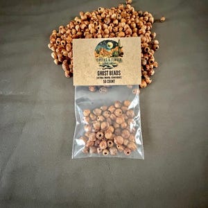 May include: A clear plastic bag containing small, round, light brown natural juniper cedar beads, with a tan cardboard header card labeled Cactus & Timber Ghost Beads Natural Juniper Cedar Beads 50 Count, surrounded by a loose pile of identical beads.