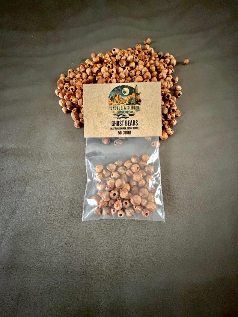 May include: A clear plastic bag containing small, round, light brown natural juniper cedar beads, with a tan cardboard header card labeled Cactus & Timber Ghost Beads Natural Juniper Cedar Beads 50 Count, surrounded by a loose pile of identical beads.